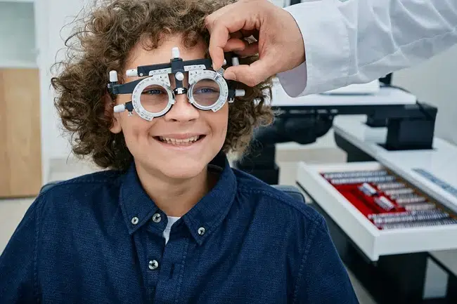 miopia_infantil_evaluacion_visual_ocumed_queretaro_650x433 Menor durante una evaluación visual mientras un profesional ajusta un armazón de prueba para revisar su visión y graduación.