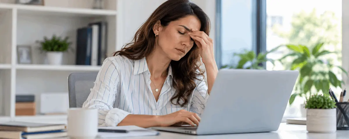 Mujer con los ojos cerrados y una mano sobre el puente de la nariz mientras trabaja frente a una computadora portátil, ilustrando fatiga visual.