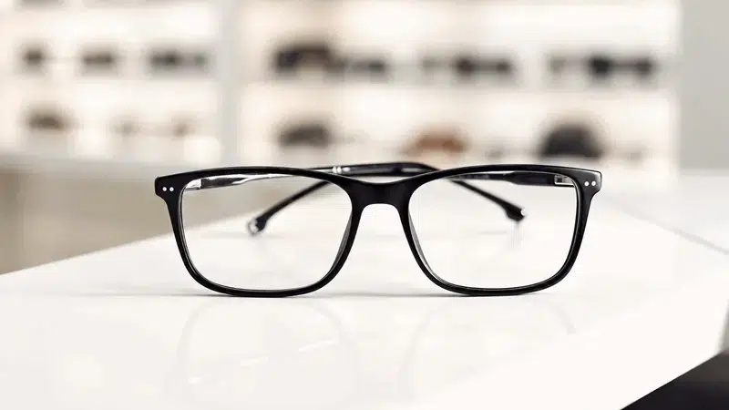 Lentes oftálmicos de armazón negro rectangular sobre una superficie blanca en una óptica, imagen ilustrativa de corrección visual para hipermetropía.